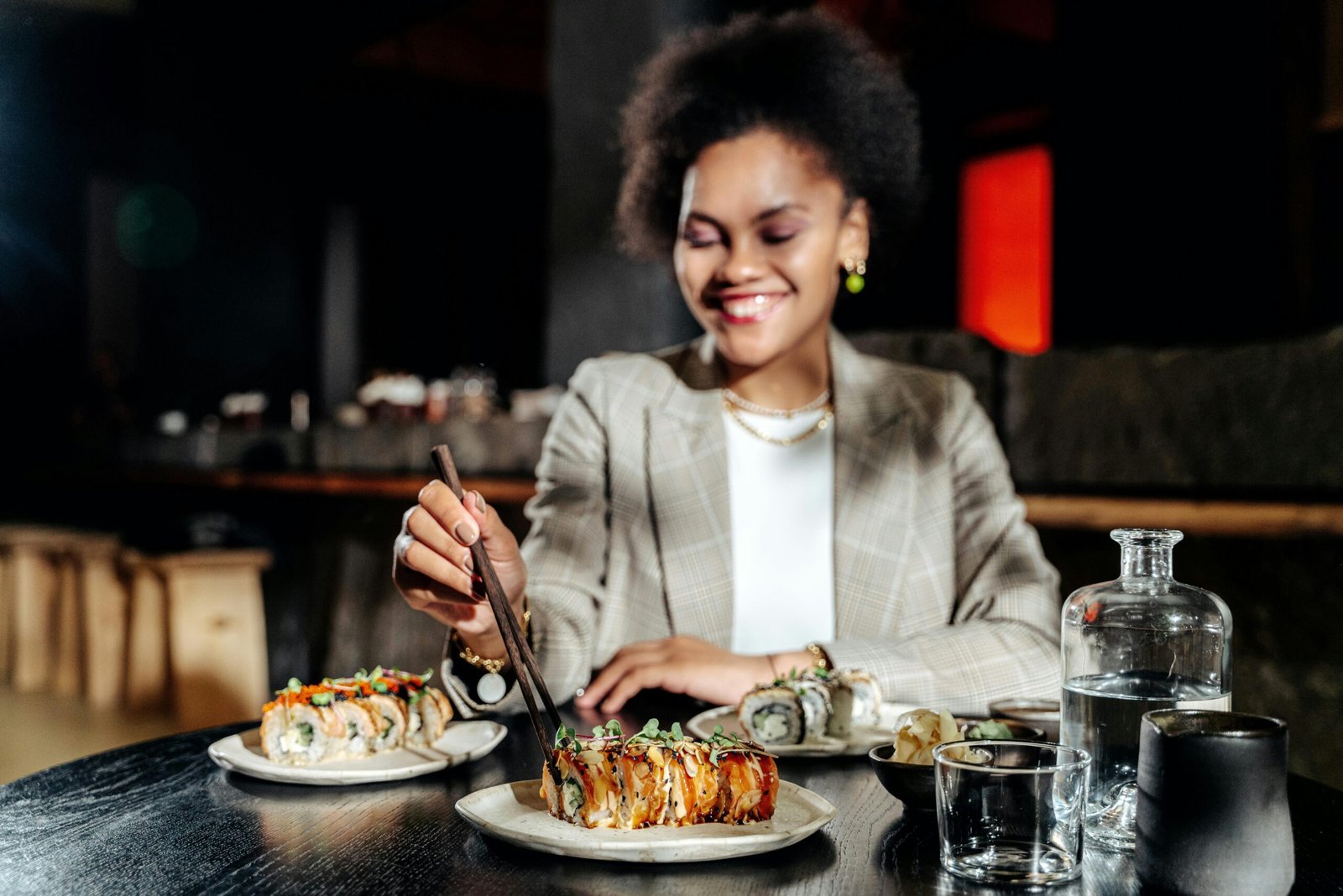 A smiling woman enjoys sushi with chopsticks at a cozy restaurant.